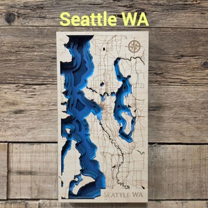 May include: A rectangular wooden bathymetric map of Seattle WA featuring laser-cut layers of light-colored wood. The water areas are depicted in deep blue, showing varying depths. A compass rose is engraved in the top right corner.