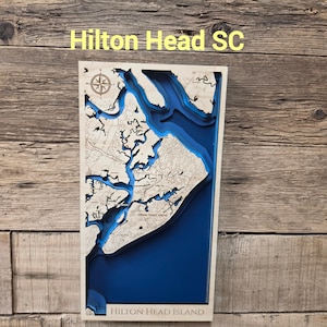 May include: A rectangular wooden bathymetric map of Hilton Head, South Carolina, featuring layered light wood landmasses and deep blue water depths. The map includes a compass rose, intricate coastline details, and the text Hilton Head Island.