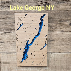 May include: A rectangular wooden wall map of Lake George NY featuring a laser-cut design with deep blue water layers set into the light-colored wood. The map includes a compass rose in the upper left corner and the text Lake George NY at the bottom.