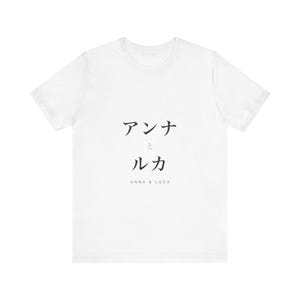 以下が含まれることがあります： 中央の胸元に「アンナとルカ」という黒い日本語のテキストと、そのすぐ下に小さなフォントで「ANNA & LUCA」という英語のテキストがプリントされた、白い半袖のTシャツです。