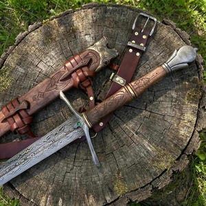 Könnte beinhalten: Ein Schwert mit gemusterter Metallklinge, einem geschnitzten Holzgriff und einem Metallknauf. Ein grüner Stein ist am Parier eingesetzt. Das Schwert wird von einer braunen Lederscheide mit Messingbeschlägen und einem Ledergürtel begleitet.