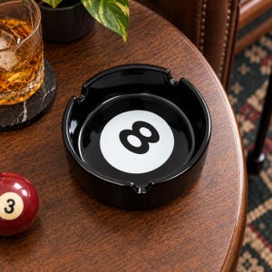 May include: A round black ashtray featuring a white circular centre with a black number eight graphic. The ashtray has four notches along the rim for resting items. The design mimics the appearance of a standard eight ball from a pool set.