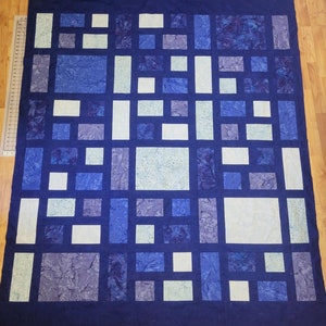 Puede incluir: Una colcha con un patrón geométrico de cuadrados y rectángulos en tonos de azul y blanco. La colcha está hecha de tela con un efecto jaspeado o tie-dye.