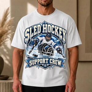 Könnte beinhalten: Ein weißes T-Shirt mit einer Grafik eines Schlittenhockeyspielers mit Helm und Trikot, der Schläger auf dem Eis hält. Das Design enthält den Text SLED HOCKEY und SUPPORT CREW in blauer und weißer Schrift mit eisigen, bergigen Akzenten.