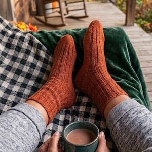 May include: A pair of burnt orange knitted socks with a vertical ribbed texture, worn on feet resting on a black and white checkered blanket, with a dark teal ceramic mug filled with a brown beverage held in the foreground.
