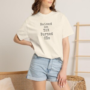 May include: A cream-colored short-sleeve t-shirt featuring black typewriter-style text on the front that reads: Raised on Y2K Burned CDs. The shirt has a crew neckline and a relaxed fit, paired with light blue denim shorts with a cuffed hem.