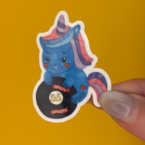 May include: A blue unicorn sticker with a rainbow mane and horn, holding a black vinyl record. The record has the number 55 in the center, with the words "Spicy" and "Smooth" on the top and bottom. The sticker has the name "Thea" on the side.
