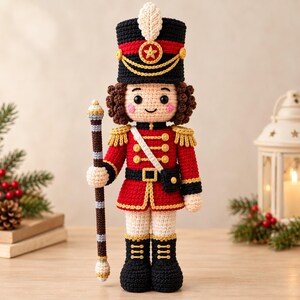 May include: A crocheted nutcracker doll wearing a red and black uniform with gold trim, a tall black hat with a white feather, and brown curly hair. The doll holds a tall, thin, brown and white striped staff and features a smiling face.