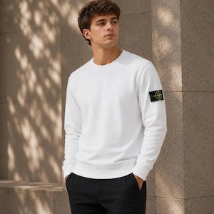 May include: A solid white long-sleeve crewneck sweatshirt featuring a black, yellow, and green compass-style patch logo on the upper left sleeve, paired with plain black trousers. The garment has a ribbed neckline, cuffs, and hem.