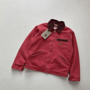 May include: A red canvas jacket featuring a dark brown corduroy collar, a front zipper closure, two slanted hand pockets, and a zippered chest pocket. A square Carhartt brand patch is visible on the chest, and a paper tag is attached to the collar.