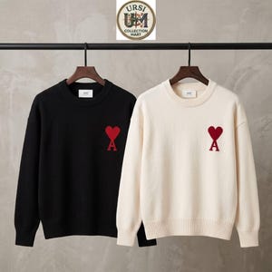 Könnte beinhalten: Zwei langärmelige Pullover mit Rundhalsausschnitt hängen an Holzkleiderbügeln. Ein Pullover ist einfarbig schwarz, der andere einfarbig cremeweiß. Beide haben ein rotes gesticktes Herz mit einem großen A auf der linken Brustseite.