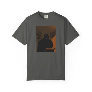 May include: A charcoal grey short-sleeve t-shirt featuring a graphic print of a dark, caped figure with pointed ears reading a book in a library setting. The illustration uses shades of black, brown, and orange, with white eyes visible on the figure.