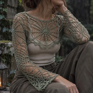 May include: A sage green long-sleeve crochet top featuring an intricate spiderweb pattern across the chest and shoulders, with flared sleeves and a round neckline, worn over a solid beige tank top and paired with loose-fitting olive green trousers.