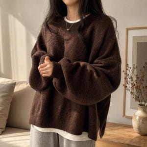 Puede incluir: Un suéter de manga larga en color marrón oscuro con cuello redondo y puños acanalados, superpuesto sobre una camiseta blanca. El suéter presenta un corte holgado y oversize con dobladillo recto y abertura lateral, combinado con pantalones grises.