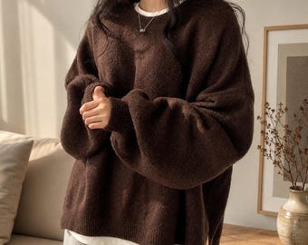 May include: A dark brown long-sleeved sweater with a crew neckline and ribbed cuffs, layered over a white undershirt. The sweater features a loose, oversized fit with a straight hemline and a side slit, paired with grey trousers.