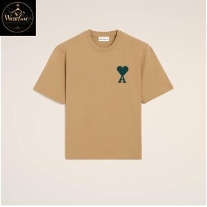 May include: A tan short-sleeved t-shirt with a crew neckline. The chest features a dark green pixelated heart and letter A logo. The garment has a straight hem and a white interior neck label with the text AMI PARIS printed in black lettering.