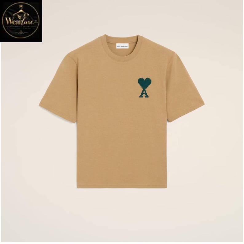 May include: A tan short-sleeved t-shirt with a crew neckline. The chest features a dark green pixelated heart and letter A logo. The garment has a straight hem and a white interior neck label with the text AMI PARIS printed in black lettering.