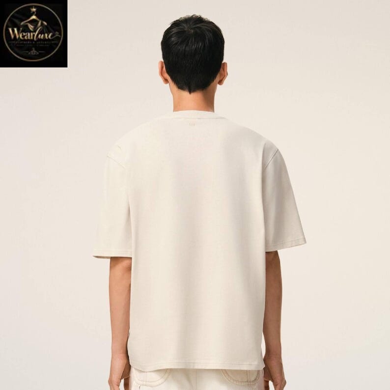 May include: A plain, short-sleeved, off-white t-shirt with a crew neckline. The garment is shown from the back, featuring a solid, unpatterned fabric construction with a standard fit, clean hemline, and simple stitching along the sleeves and collar.