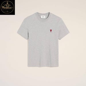 May include: A light grey short-sleeved t-shirt featuring a crew neckline and a small red heart-shaped logo embroidered on the left chest. The shirt includes a white interior neck label with the text ami printed in black lettering.
