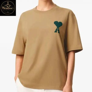 May include: A tan short-sleeved t-shirt featuring a dark green embroidered heart-shaped logo with a letter A integrated into the design on the upper left chest. The shirt has a crew neckline and a relaxed fit, providing a simple and clean look.