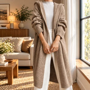 May include: A long, beige, open-front hooded cardigan made of a soft knit fabric. The cardigan features long sleeves with ribbed cuffs and a straight hemline. It is worn over a white mock-neck top and white trousers, creating a layered look.