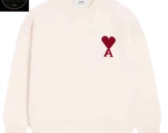 May include: A cream-colored long-sleeve crewneck sweater featuring a red embroidered heart and letter A logo on the left chest. The garment has a ribbed neckline, cuffs, and hem, with a small white brand label visible inside the collar area.