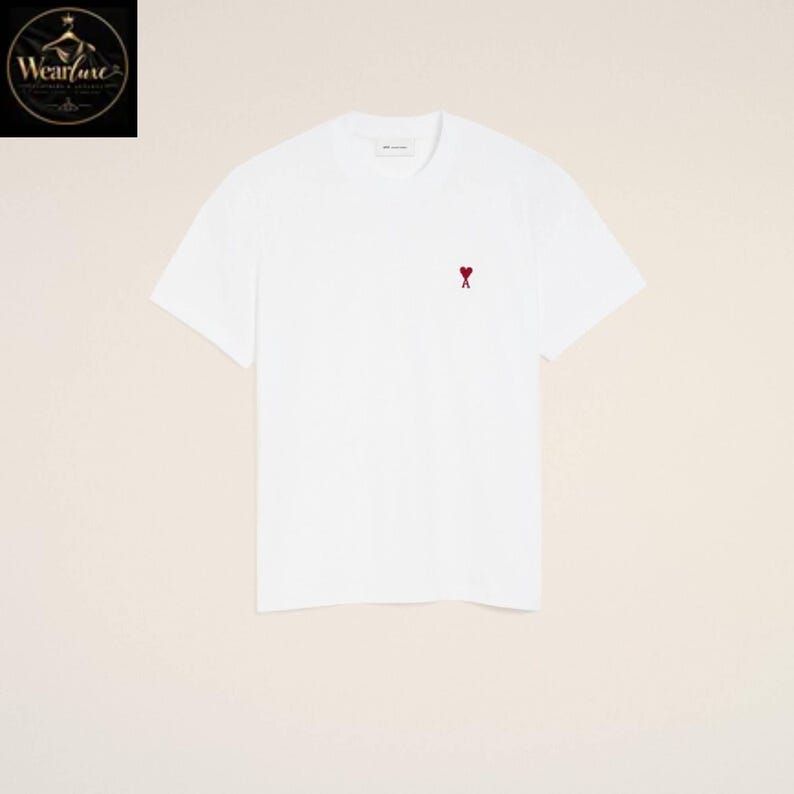 May include: A plain white short-sleeved t-shirt featuring a small, embroidered red heart logo with a cross symbol centered on the upper left chest area. The garment has a classic crew neckline and a clean, solid white fabric construction throughout.