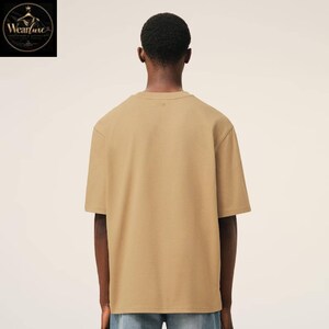 May include: A solid tan short-sleeved t-shirt with a crew neckline, shown from the back. The garment features a plain, unpatterned design with a standard fit, short sleeves, and a straight hemline, constructed from a smooth, opaque fabric.