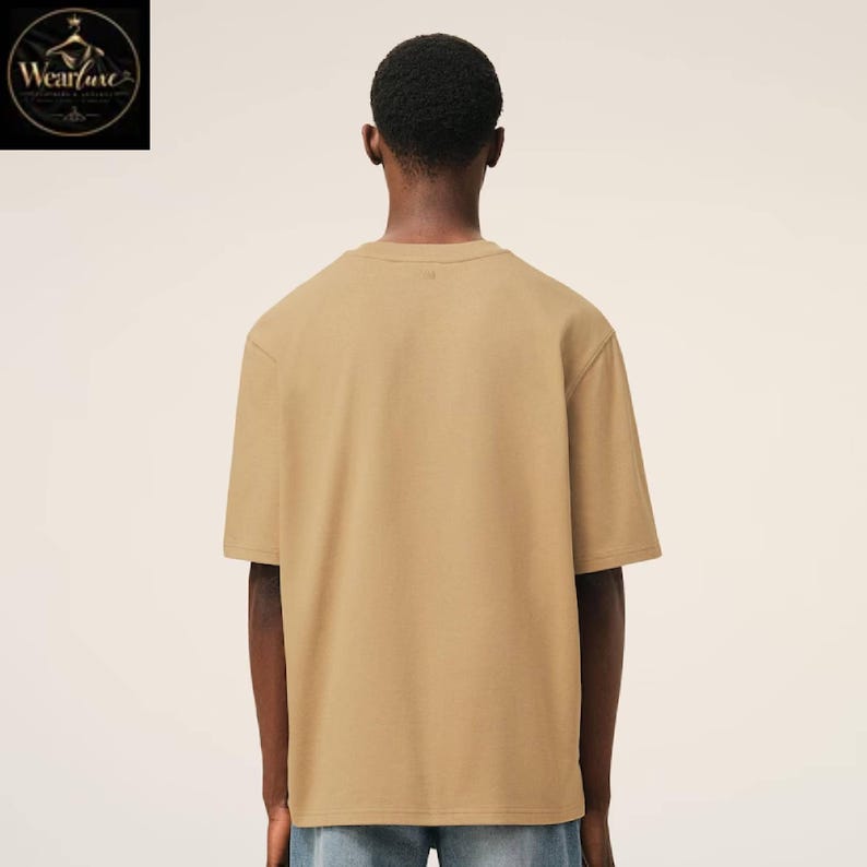 May include: A solid tan short-sleeved t-shirt with a crew neckline, shown from the back. The garment features a plain, unpatterned design with a standard fit, short sleeves, and a straight hemline, constructed from a smooth, opaque fabric.