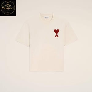 May include: A short-sleeved, off-white crew neck t-shirt featuring a small, pixelated dark red heart icon with the letter A integrated at the center of the chest. The garment has a simple, clean design with a visible brand label at the inner neckline.
