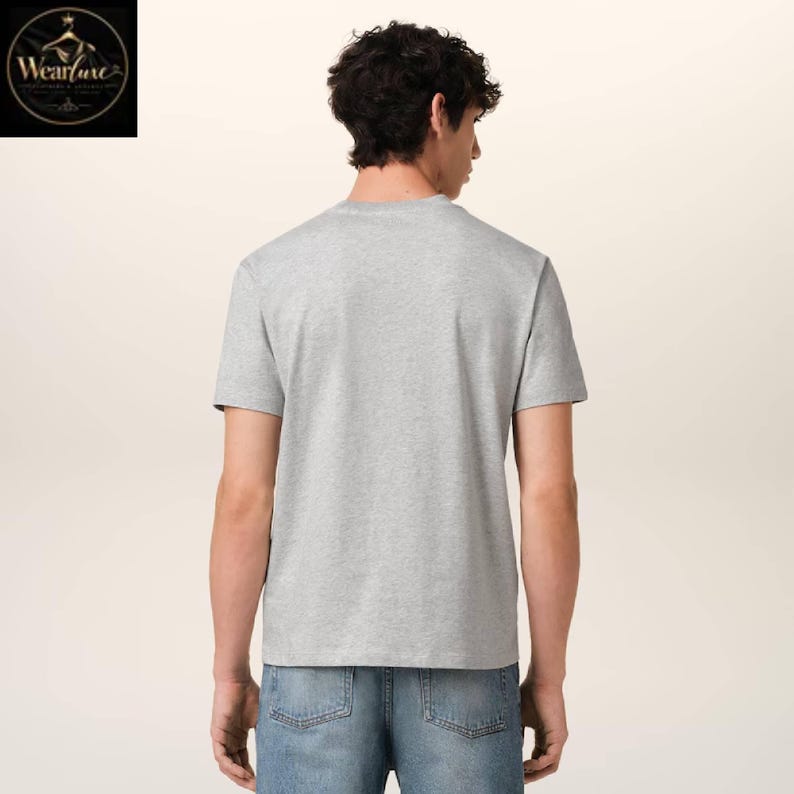 May include: A short-sleeved heather gray t-shirt with a crew neckline, worn with blue denim jeans. The view shows the back of the garment, which features a plain, solid-colored fabric design. The shirt is paired with classic blue denim pants.
