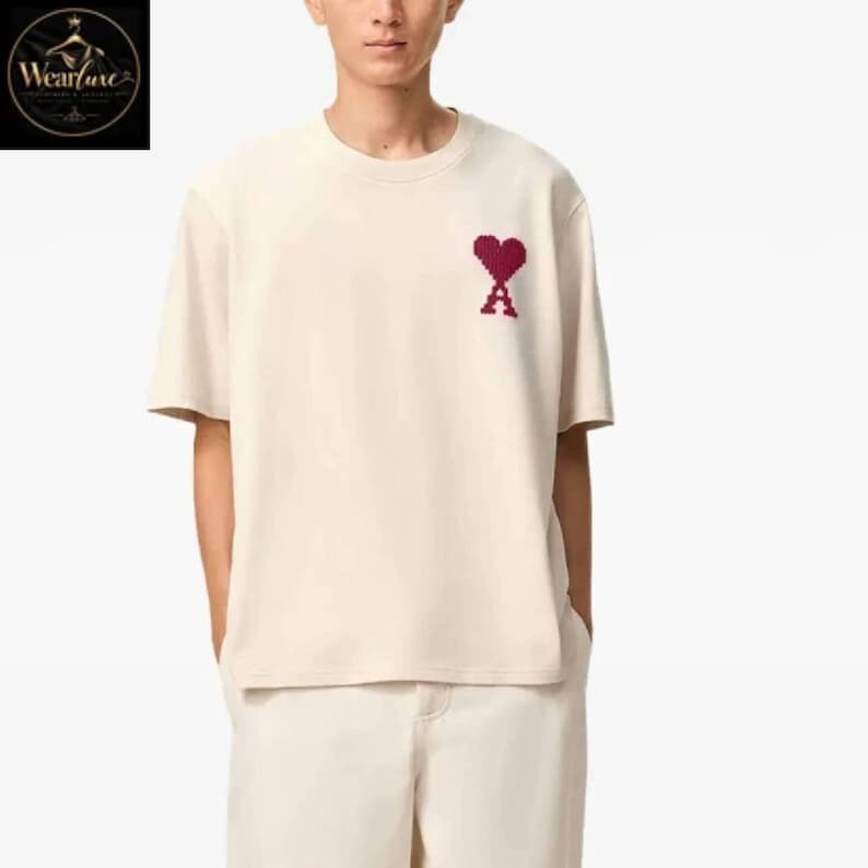 May include: A short-sleeved, crew-neck t-shirt in a solid off-white color. The garment features a small, embroidered pixelated heart logo in a deep red shade positioned on the upper left chest area. The fabric appears to be a smooth cotton knit.