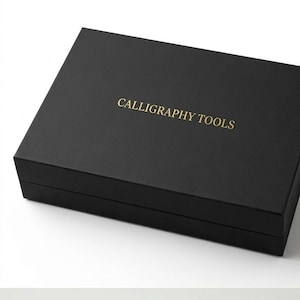 Puede incluir: Una caja de almacenamiento rectangular de color negro con acabado mate. La tapa presenta el texto CALLIGRAPHY TOOLS impreso en letras mayúsculas doradas centradas en la parte superior. La caja está cerrada y tiene un diseño estructurado.