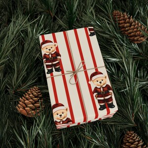 Könnte beinhalten: Ein rechteckiges Geschenkpaket, eingewickelt in cremefarbenes Papier mit vertikalen roten Streifen und einem wiederkehrenden Muster aus Teddybären, die rote Weihnachtsmannanzüge und Mützen tragen. Das Paket ist mit einer dünnen braunen Schnur umwickelt.