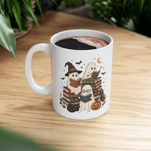 Könnte beinhalten: Ein weißer Keramikbecher mit einer farbenfrohen Illustration von drei Geistern, die auf einem Bücherstapel sitzen und lesen. Das Motiv zeigt zudem eine schwarze Katze, einen Kürbis, eine Kerze, Fledermäuse und eine Mondsichel im Cartoon-Stil.
