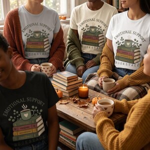 May include: A group of people wearing t-shirts with a graphic print of a stack of books topped with a steaming mug and the text "EMOTIONAL SUPPORT BOOKSTACK". The shirts are white, light blue, and black, paired with open-front knit cardigans.