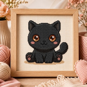 Puede incluir: Un marco de madera cuadrado que contiene un bordado de punto de cruz de un gato negro con grandes ojos color ámbar, mejillas rosadas y almohadillas rosadas. El gato está sentado sobre un fondo de tela beige claro con una cuadrícula visible.