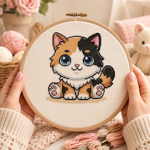 Puede incluir: Un bastidor de bordado circular de madera que contiene un patrón de punto de cruz de un gatito calicó. El gatito tiene grandes ojos azules, manchas negras y naranjas, y almohadillas rosas en las patas sobre un fondo de tela blanca.