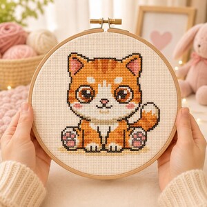 Puede incluir: Un bastidor de bordado circular de madera que contiene un patrón de punto de cruz de un gato atigrado naranja sentado con ojos grandes y oscuros, orejas rosas y marcas blancas en la cara y el pecho, sobre una tela de color crema.