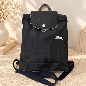 Könnte beinhalten: Ein schwarzer Nylon-Rucksack mit einer strukturierten schwarzen Klappe und einem weißen Druckknopfverschluss. Der Rucksack verfügt über einen in Plastik eingewickelten Tragegriff, verstellbare schwarze Schultergurte und ein weißes Pferdelogo.