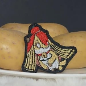 Könnte beinhalten: Ein gestickter Aufnäher mit einer stilisierten Zeichentrickfigur mit langem blondem Haar, einem roten Stirnband und einem weißen Outfit, umrandet mit schwarzem Garn. Der Aufnäher liegt vor drei rohen, hellbraunen Kartoffeln auf einem weißen Teller.