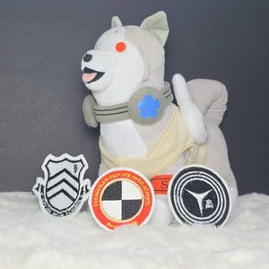 May include: A plush dog toy with grey and white fur, wearing a beige vest and a grey collar with a blue star emblem. Three circular and shield-shaped embroidered patches are placed in front, featuring black, white, red, and grey geometric designs.