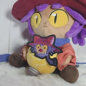 Puede incluir: Un peluche que representa un personaje con cabello morado, grandes ojos amarillos y un sombrero granate ancho. El personaje lleva una bufanda azul y una túnica rojo anaranjado, sosteniendo un objeto amarillo con forma de bombilla y un parche bordado.