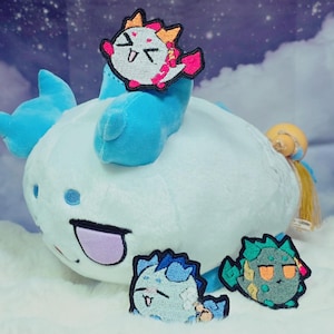 May include: A light blue plush creature with a rounded body, blue ears, and a purple mouth. It features three embroidered patches: one white and pink creature on top, and two smaller creature patches in blue and green placed in front.