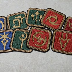 Puede incluir: Una colección de nueve parches cuadrados bordados con símbolos de color dorado sobre fondos azules, verdes y rojos. Cada parche tiene un borde grueso y oscuro y muestra un emblema estilizado único cosido con hilo dorado.