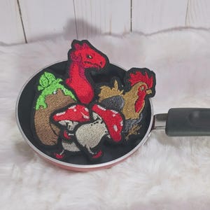May include: A black frying pan containing several embroidered patches. The patches feature a red dragon head, a brown and green tree stump, two red and white mushrooms, and a brown rooster with a red comb, all arranged inside the pan.