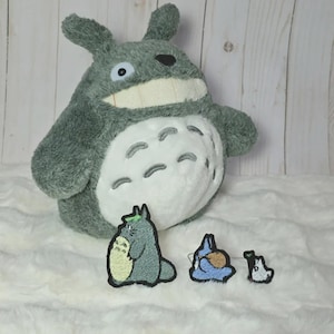 Può includere: Un peluche grigio e bianco con un corpo grande e rotondo, orecchie a punta e una bocca larga, accompagnato da tre toppe ricamate più piccole che raffigurano creature simili in grigio, blu e bianco con contorni neri e dettagli verdi.