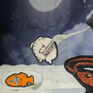 Könnte beinhalten: Ein runder, beigefarbener Stickaufnäher mit einem einfachen, liniengezeichneten Gesicht, der von einer silbernen Gabel gehalten wird. Darunter befindet sich ein kleiner weißer Teller mit einem orangefarbenen fischförmigen Aufnäher mit weißer Schleife.