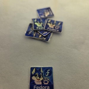 Könnte beinhalten: Eine Sammlung kleiner quadratischer Aufkleber mit einem blau-weißen Cartoon-Wolf mit Brille, einem Pfotenabdruck-Symbol, dem Buchstaben f und dem Text Fedora, der in Weiß am unteren Rand jedes Aufkleberdesigns aufgedruckt ist.