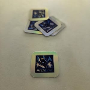 May include: A collection of square stickers featuring a holographic border and a central graphic of a stylized character with headphones next to the Arch Linux logo and the text Arch Linux printed in white and blue on a dark background.